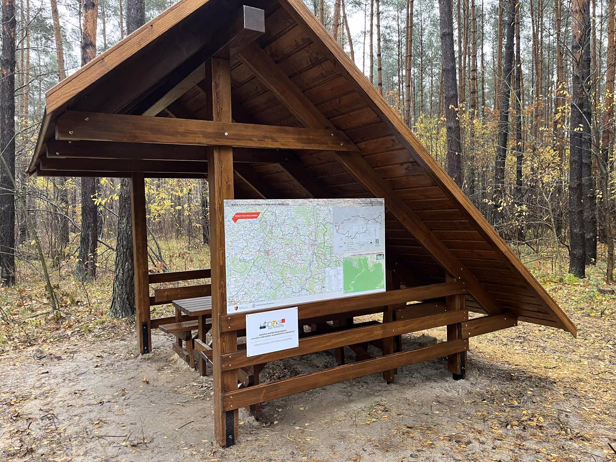 Drewniana wiata z ławkami i mapą turystyczną Powiatu Radomszczańskiego ustawiona przy Rodzinnej Ścieżce Rowerowej w otoczeniu sosnowego lasu.