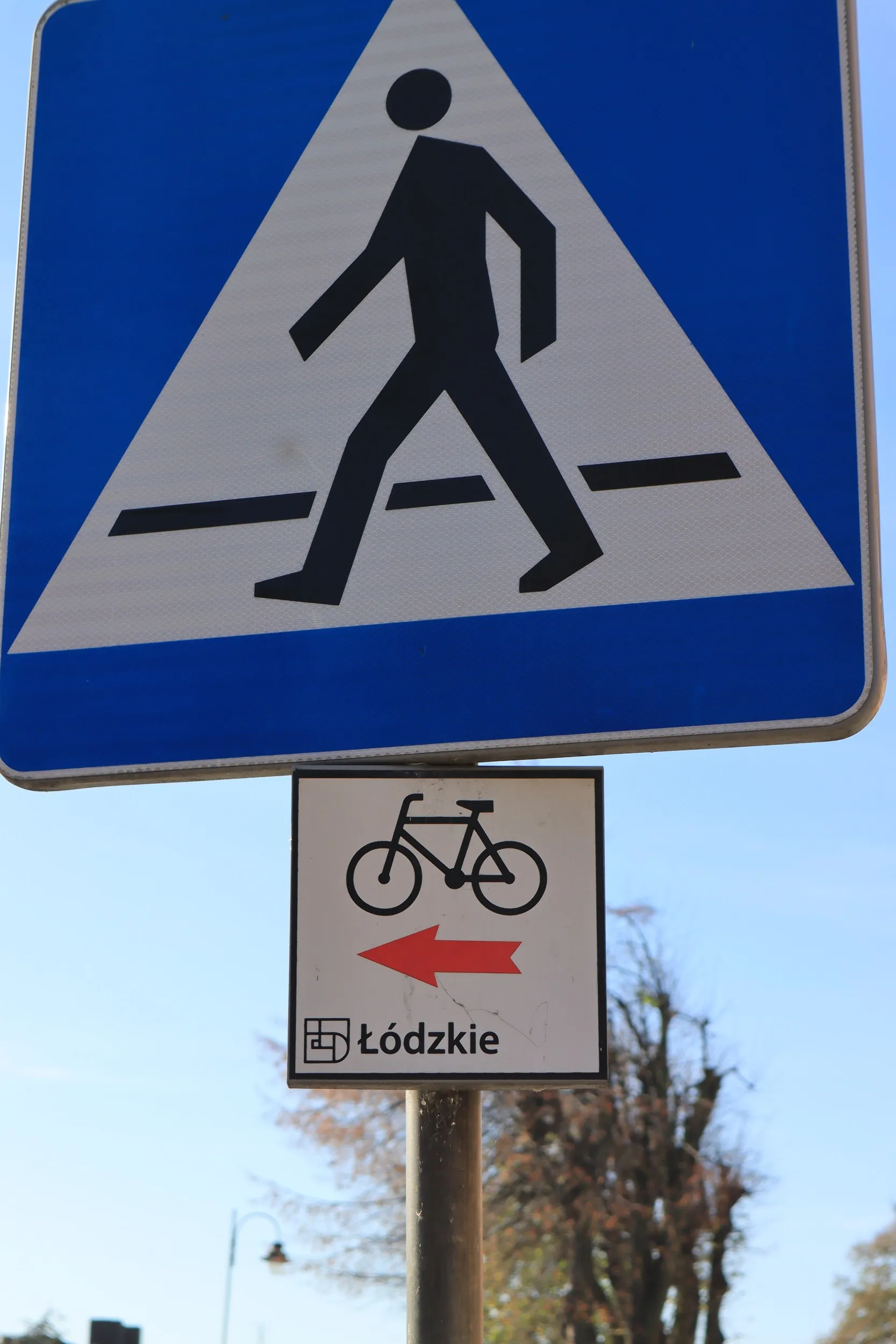 Znak przejścia dla pieszych z tabliczką wskazującą kierunek szlaku rowerowego. Oznakowanie znajduje się przy drodze w terenie zabudowanym.