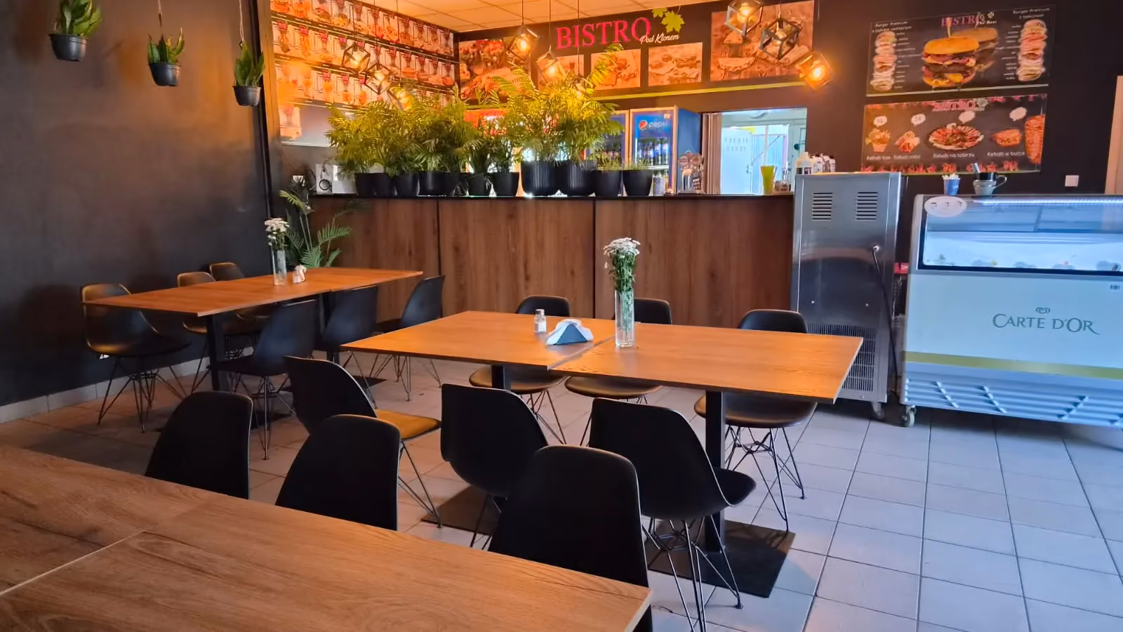 Przestrzeń konsumpcyjna Bistro Pod Klonem z komfortowym układem stolików i ofertą gastronomiczną widoczną w tle.