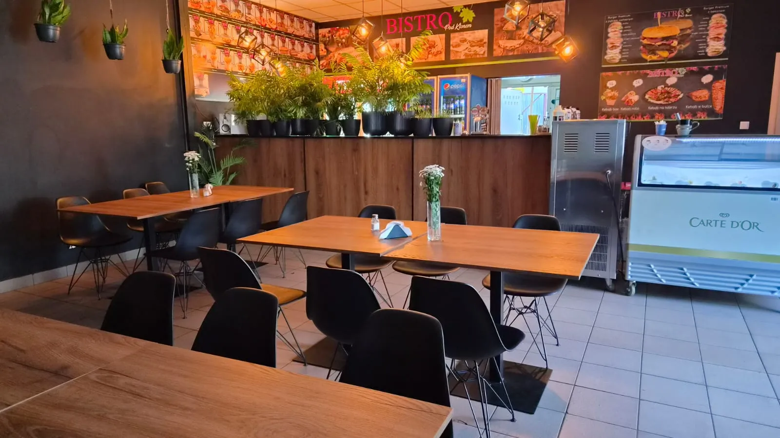 Przestrzeń konsumpcyjna Bistro Pod Klonem z komfortowym układem stolików i ofertą gastronomiczną widoczną w tle.