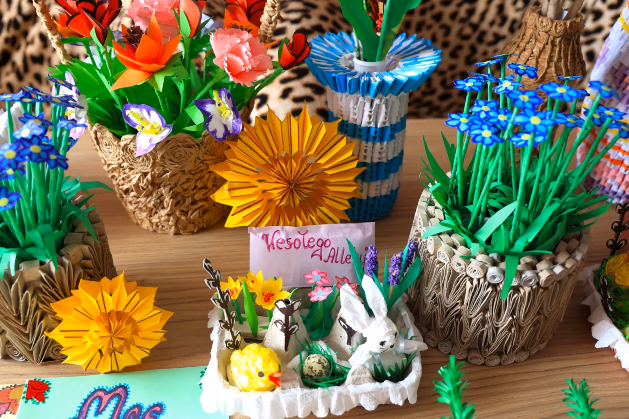 Stół zastawiony ręcznie wykonanymi dekoracjami z papieru techniką origami: kwiatami, wazonami oraz wielkanocną kompozycją z zajączkiem i kurczaczkiem
