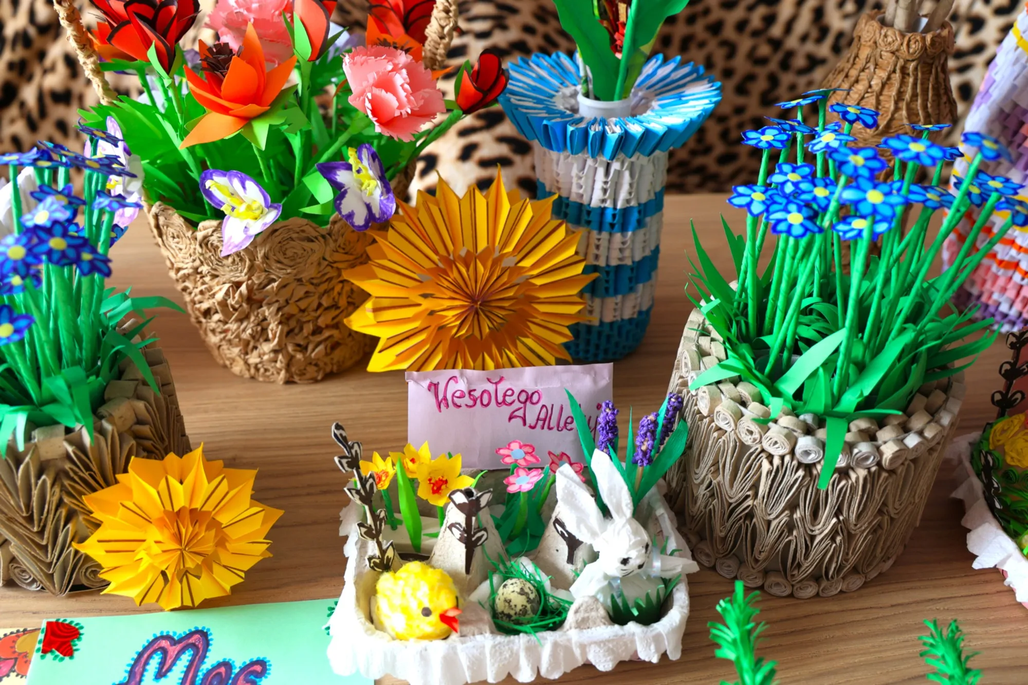Stół zastawiony ręcznie wykonanymi dekoracjami z papieru techniką origami: kwiatami, wazonami oraz wielkanocną kompozycją z zajączkiem i kurczaczkiem
