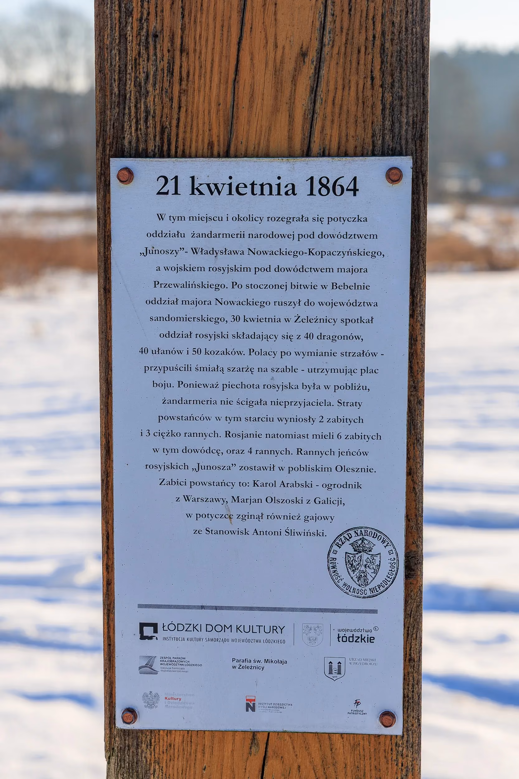 Tablica informacyjna opisująca wydarzenia z 21 kwietnia 1864 roku związane z Powstaniem Styczniowym. Tablica zamocowana jest na drewnianym słupie.