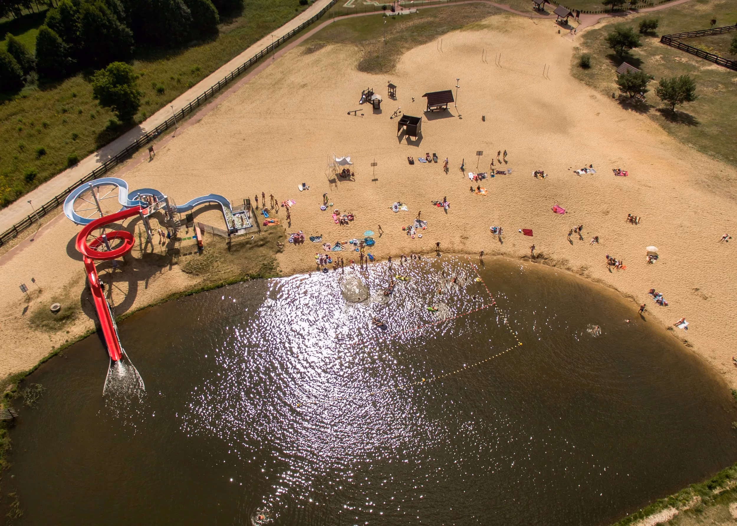 Plaża nad zalewem w Przedborzu z kąpiącymi się osobami, zjeżdżalniami wodnymi i miejscami do wypoczynku na piasku.