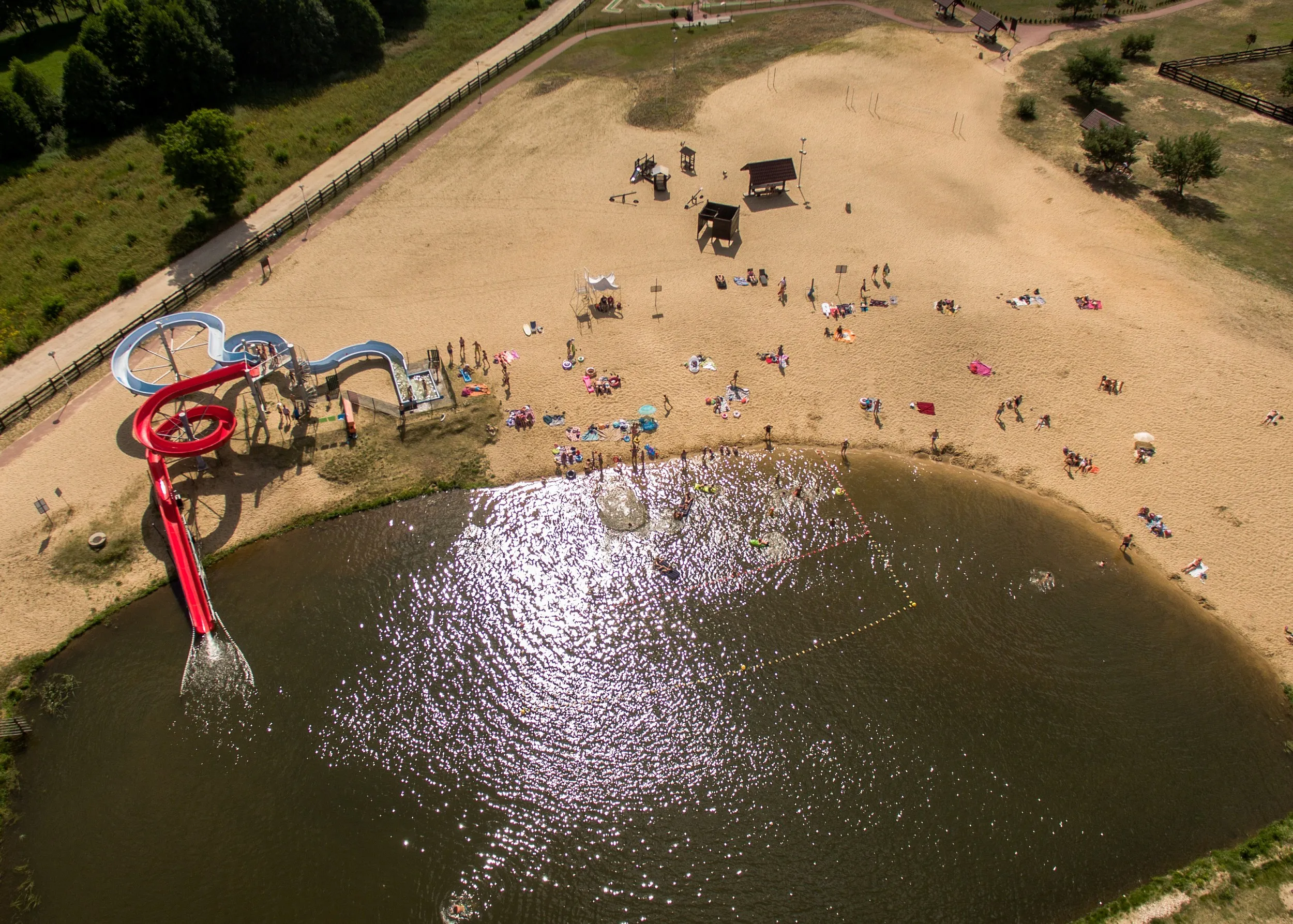 Plaża nad zalewem w Przedborzu z kąpiącymi się osobami, zjeżdżalniami wodnymi i miejscami do wypoczynku na piasku.