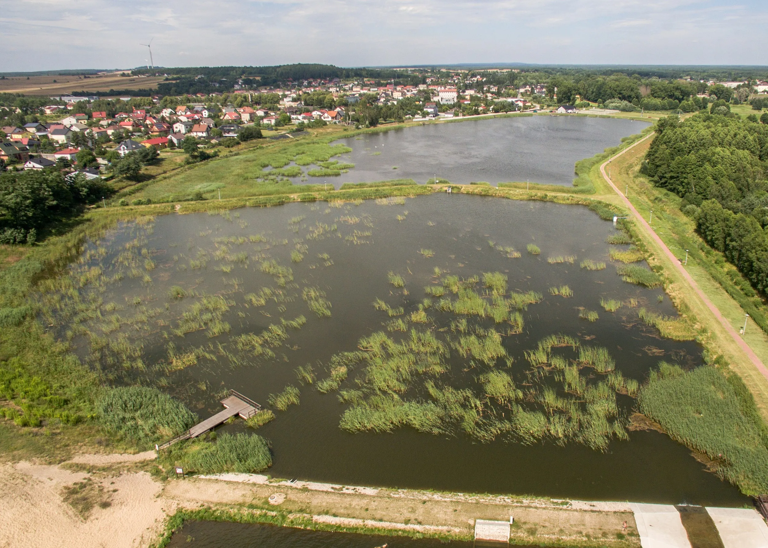 Lotniczy widok na zalew w Przedborzu, trzcinowiska, pomost, ścieżkę nad wodą oraz zabudowę miasta w oddali.