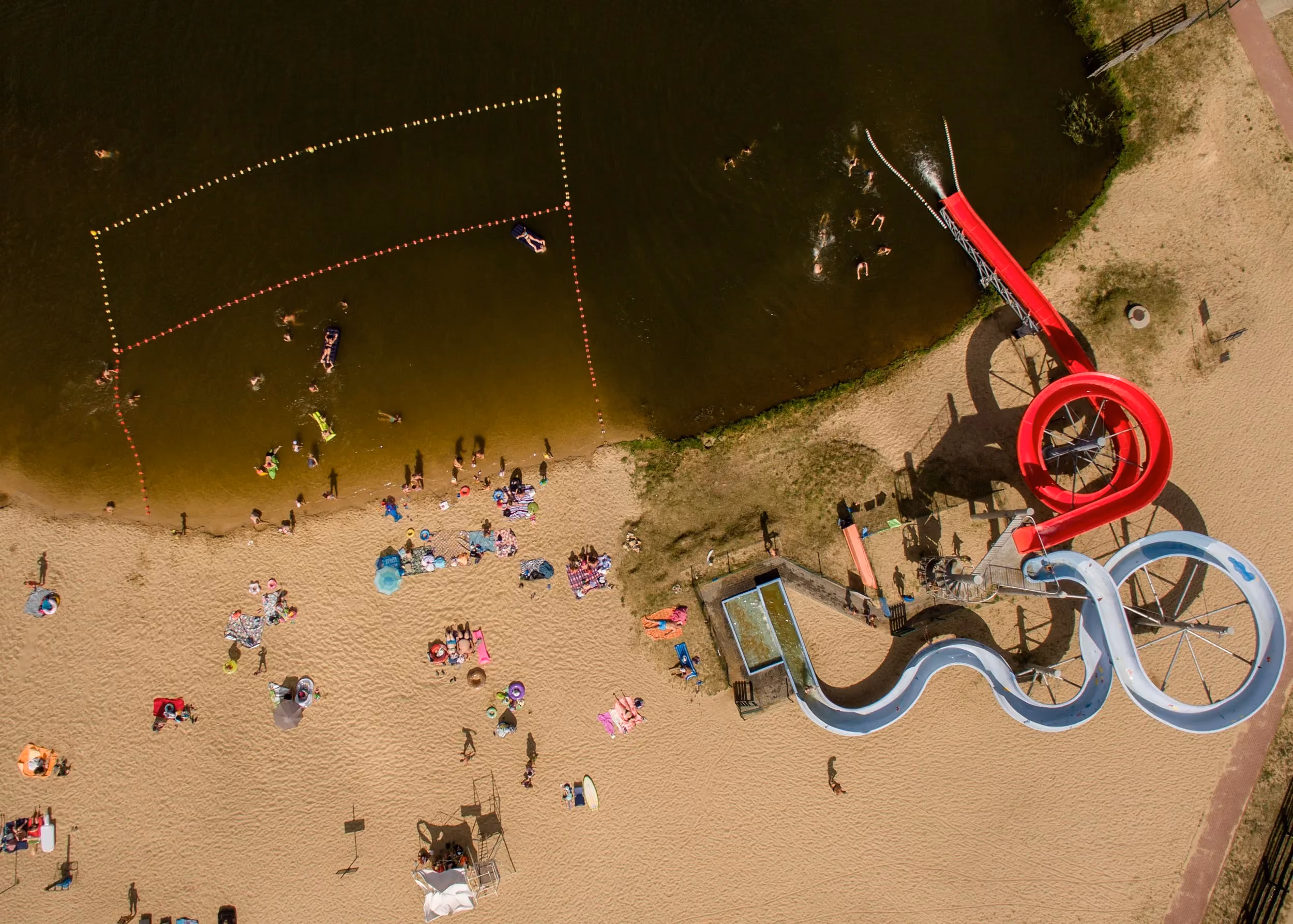 Widok z góry na kąpielisko w Przedborzu z plażą, kąpiącymi się osobami, wyznaczoną strefą pływania i zjeżdżalniami.