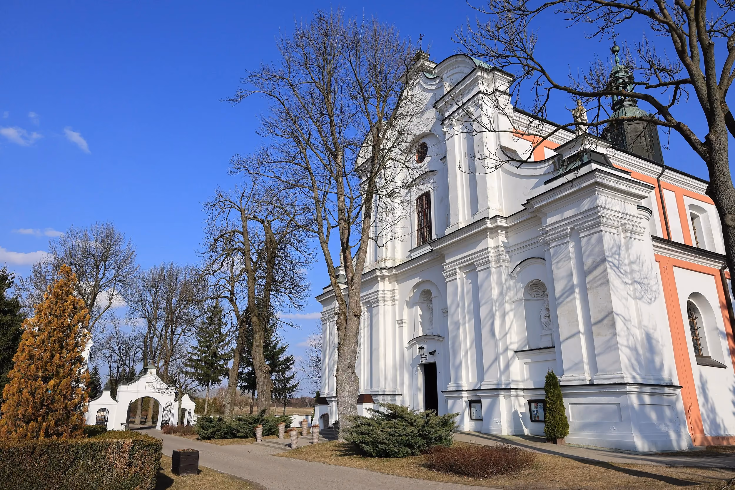 Front kościoła o barokowej architekturze, otoczony zielenią. Obok widoczna zabytkowa brama prowadząca na teren świątyni.