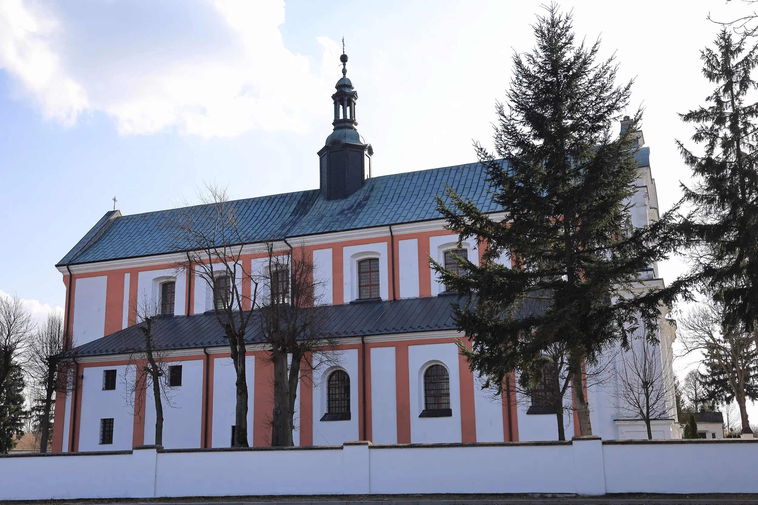 Boczna elewacja kościoła z czerwono-białą fasadą, otoczona drzewami i murem odgradzającym teren świątyni.