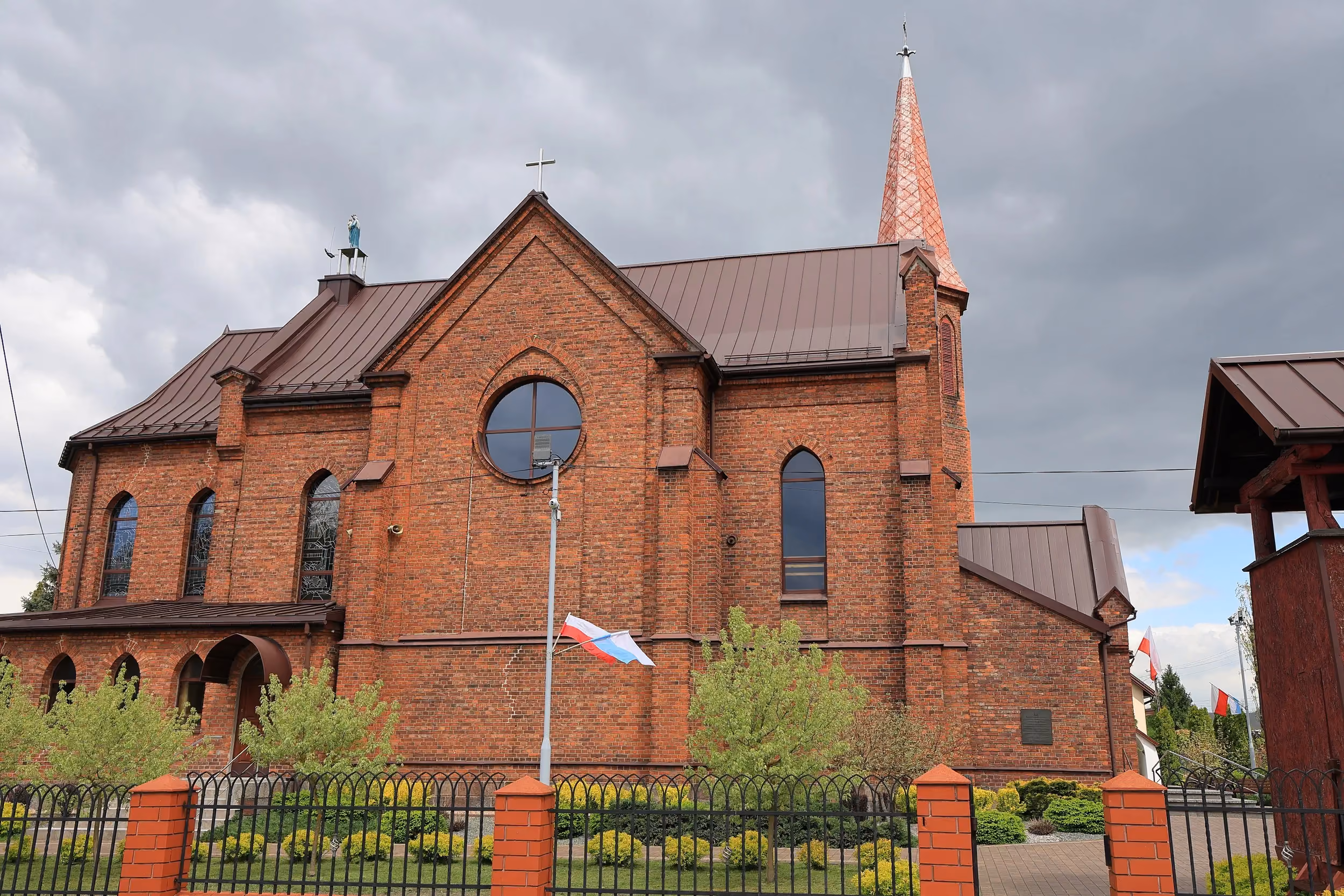Boczna elewacja ceglanego kościoła z ostrołukowymi oknami i wieżą. Przed świątynią znajdują się flagi oraz zadbana zieleń, nad budynkiem wiszą ciemne chmury.