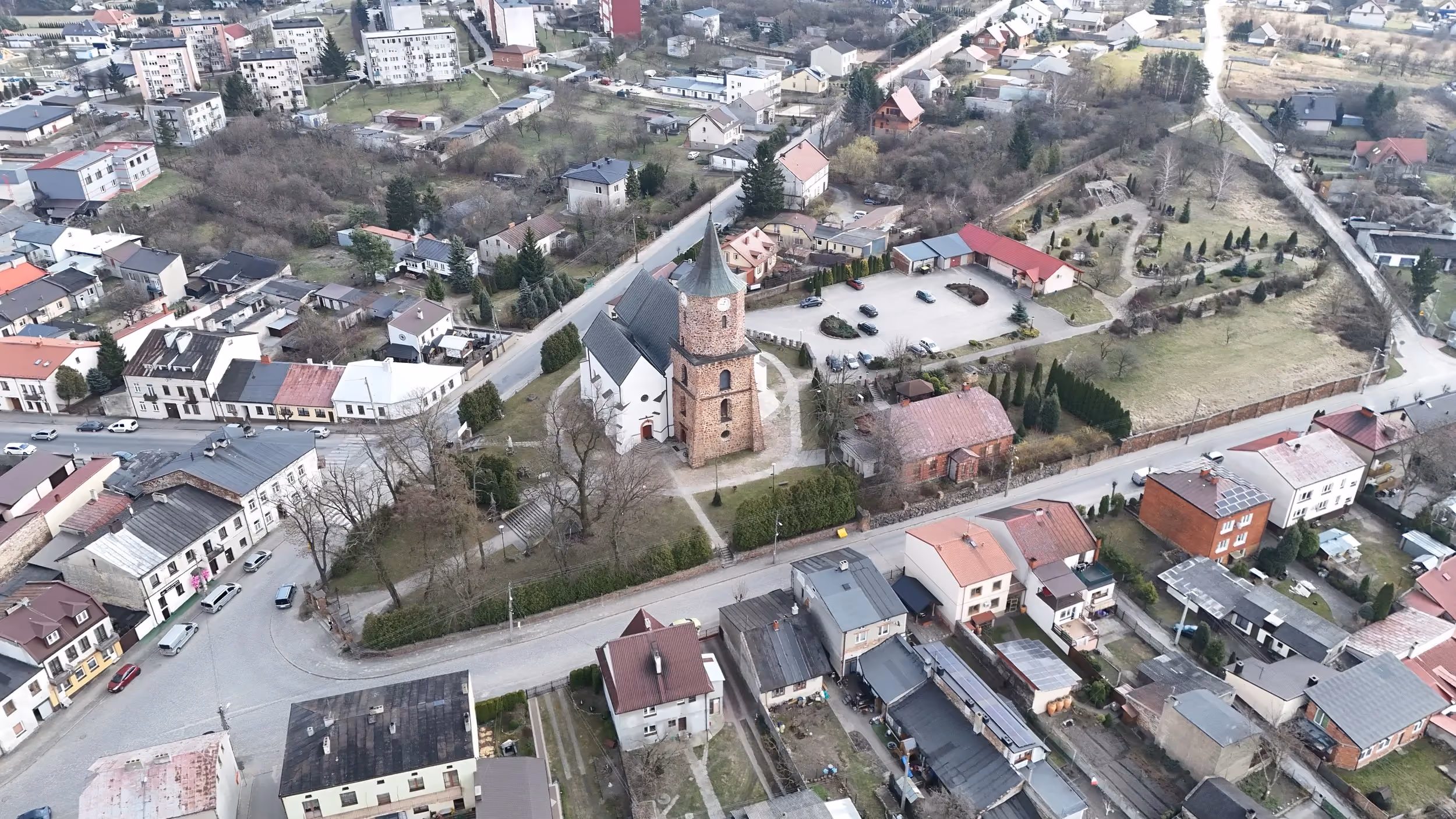 Panoramiczny widok z drona na kościół z kamienną wieżą i otaczającą zabudowę miejscowości. Wokół świątyni znajdują się domy, drogi oraz tereny zielone.
