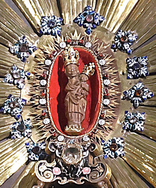 Alt: Cudowna figurka Matki Bożej z Dzieciątkiem w bogato zdobionym relikwiarzu, otoczona złotymi promieniami i dekoracyjnymi kamieniami.