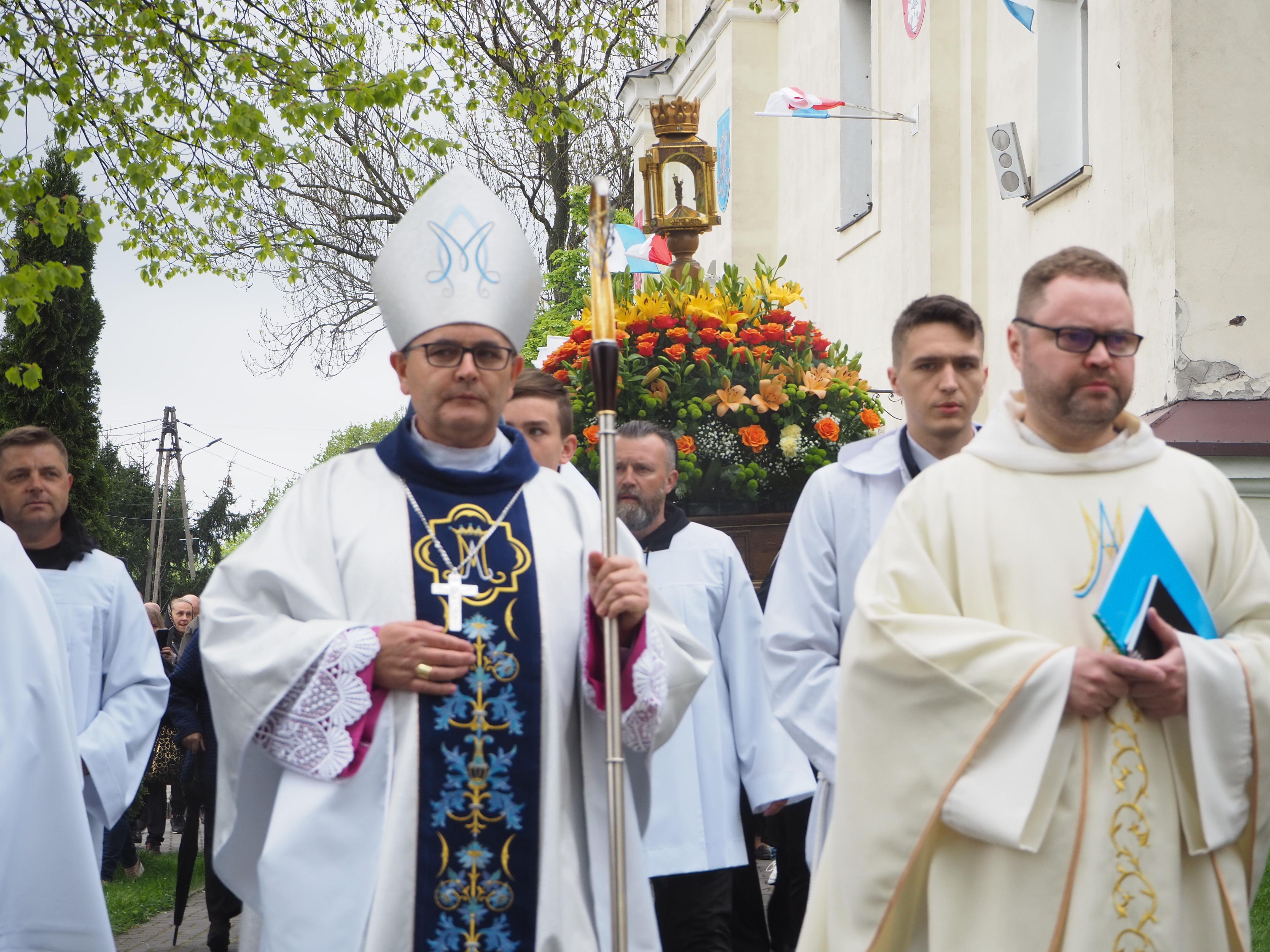 Procesja religijna w Gidlach z biskupem i duchownymi, za którymi niesiony jest relikwiarz z figurką Matki Bożej na kwiatowej dekoracji.