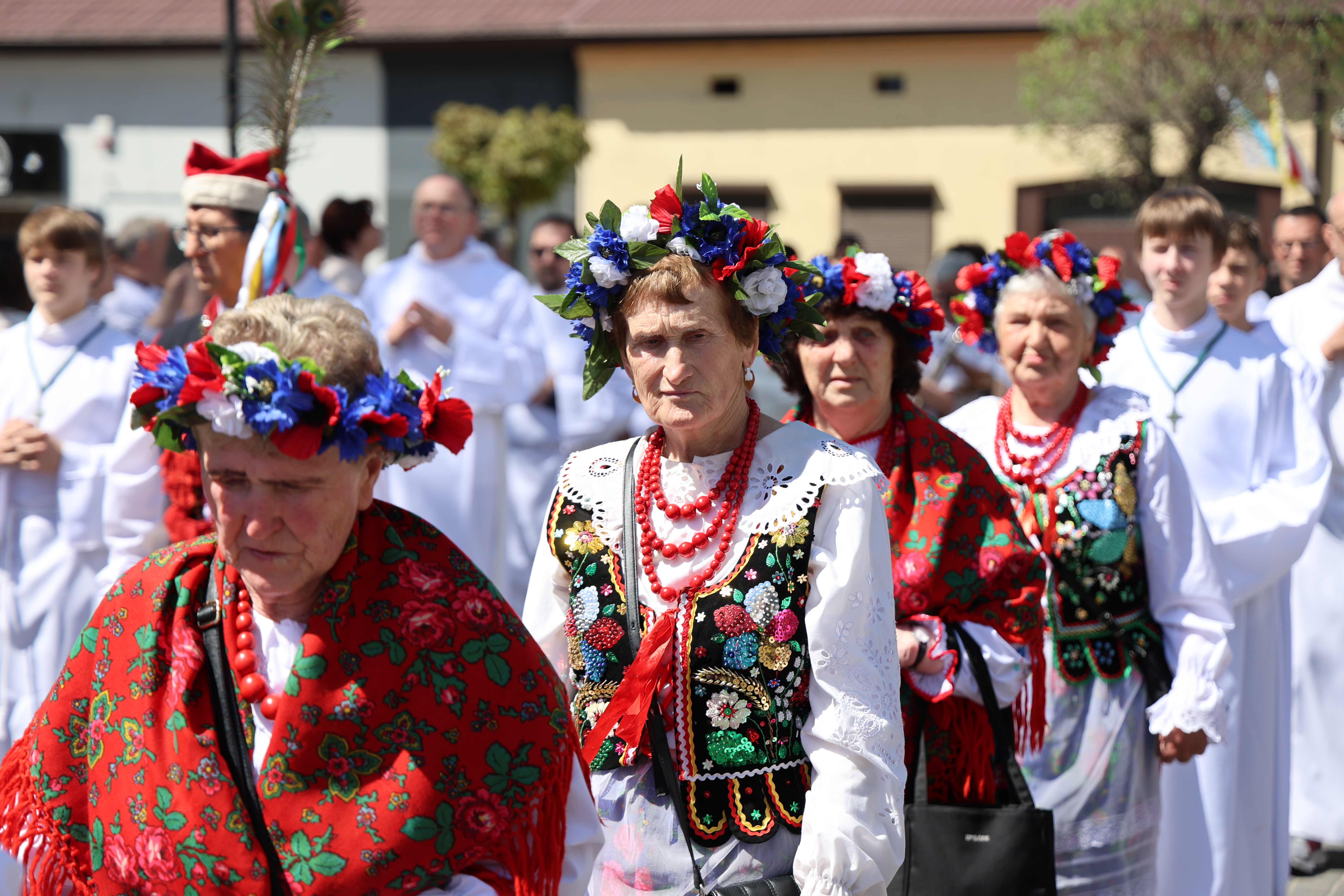 Kobiety w tradycyjnych strojach ludowych i wiankach idą w procesji religijnej w Gidlach, wśród duchownych i uczestników uroczystości.