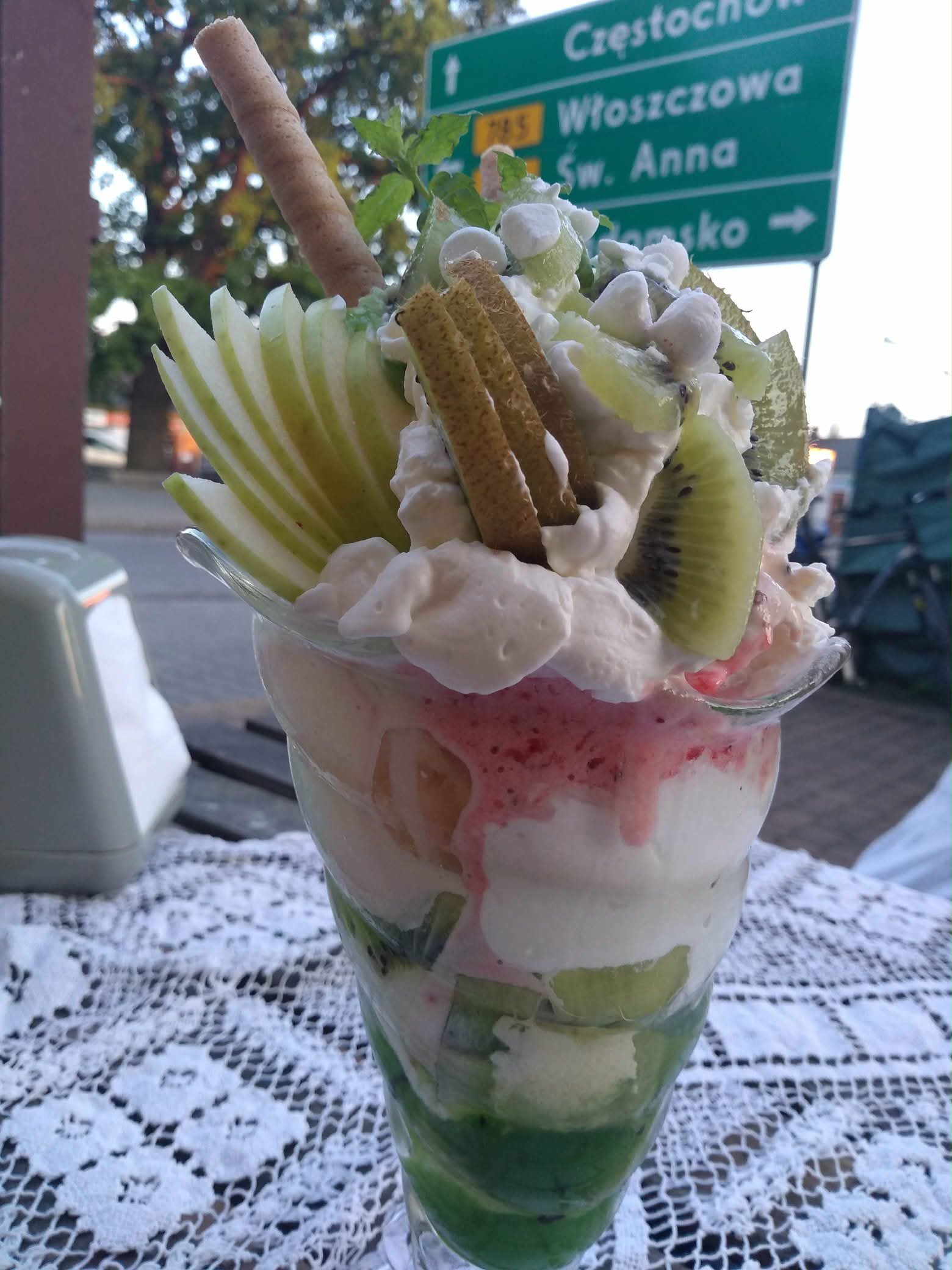 Deser lodowy z bitą śmietaną, kiwi, jabłkiem, wafelkami i piankami, podany w wysokim pucharku na stoliku z koronkowym obrusem.