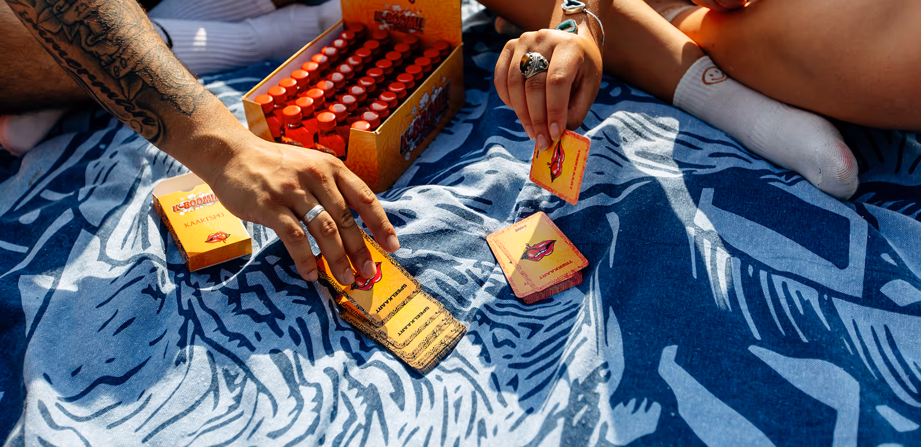 Twee personen spelen een kaartspel K-BOOM! buiten op een blauw gemêleerde deken in de zon.