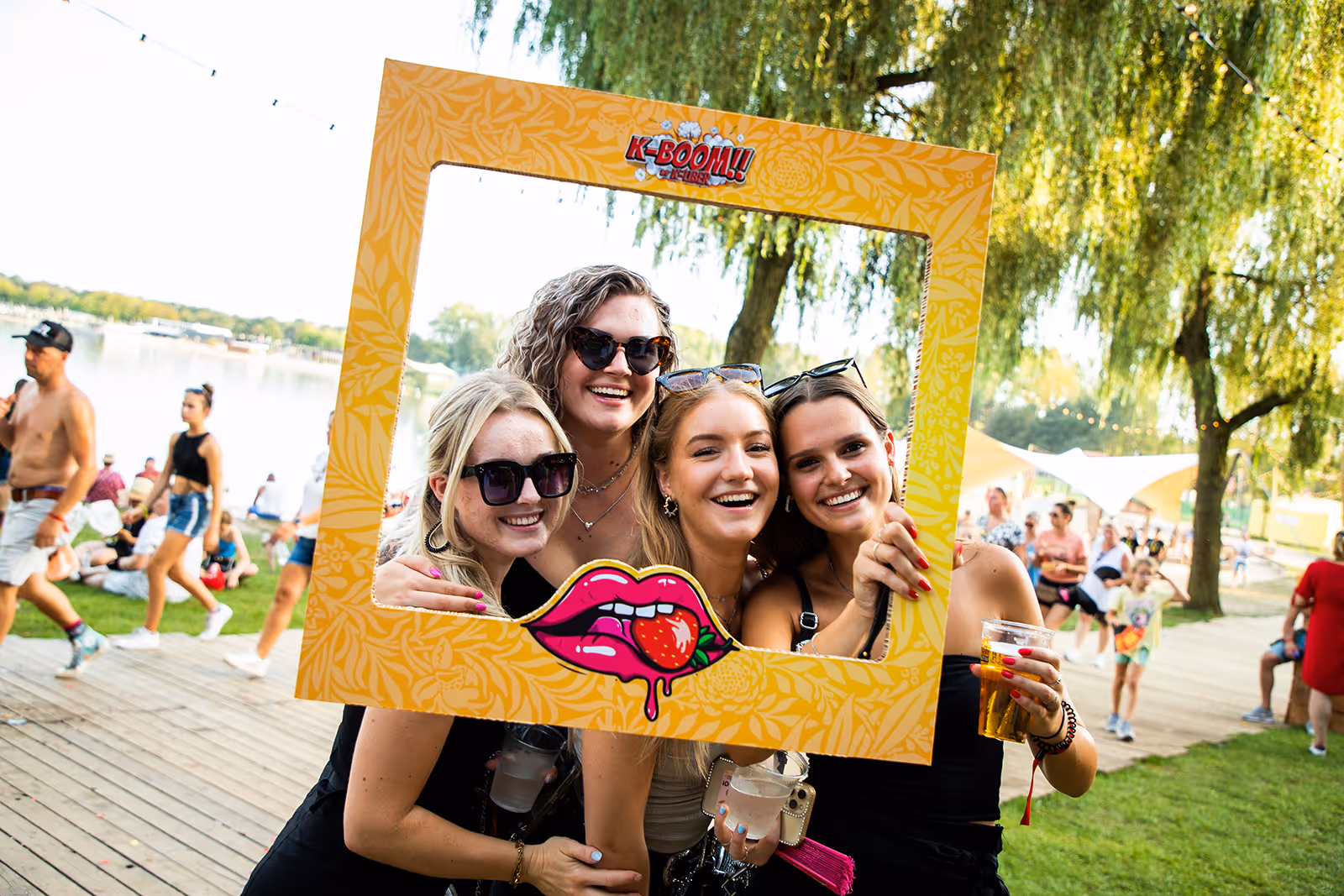 Vier lachende vrouwen poseren buiten in een gele fotolijst met een lippenstift en aardbei ontwerp tijdens een evenement bij een meer.