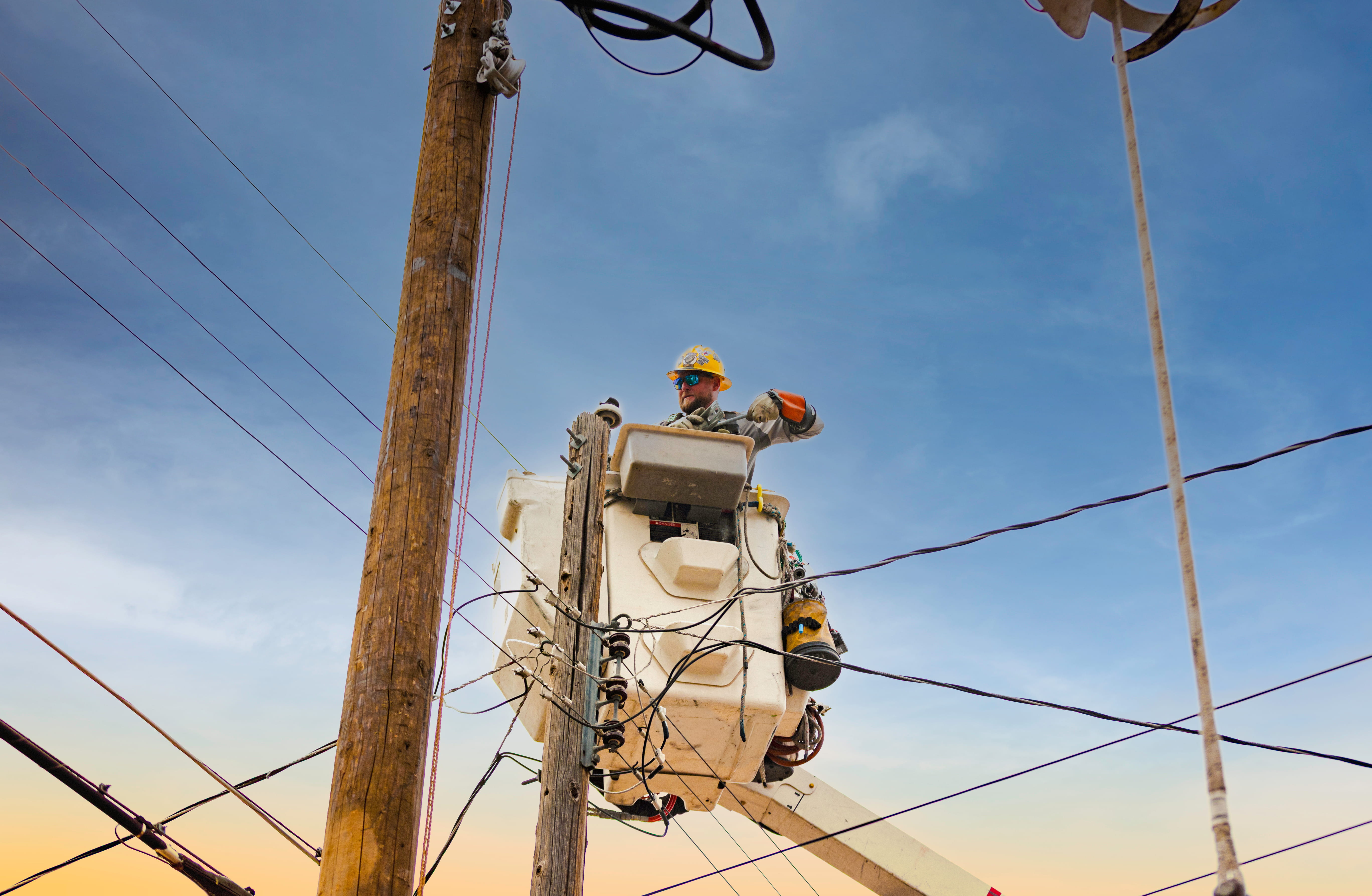 Utility worker in safety gear elevated in a bucket truck working on power lines against a clear sky.