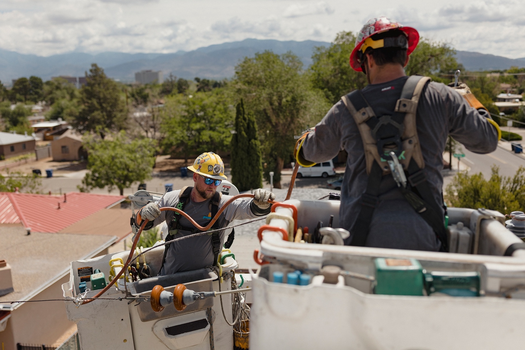 Two utility workers in safety gear handling cables from elevated bucket trucks with a suburban neighborhood and mountains in the background.