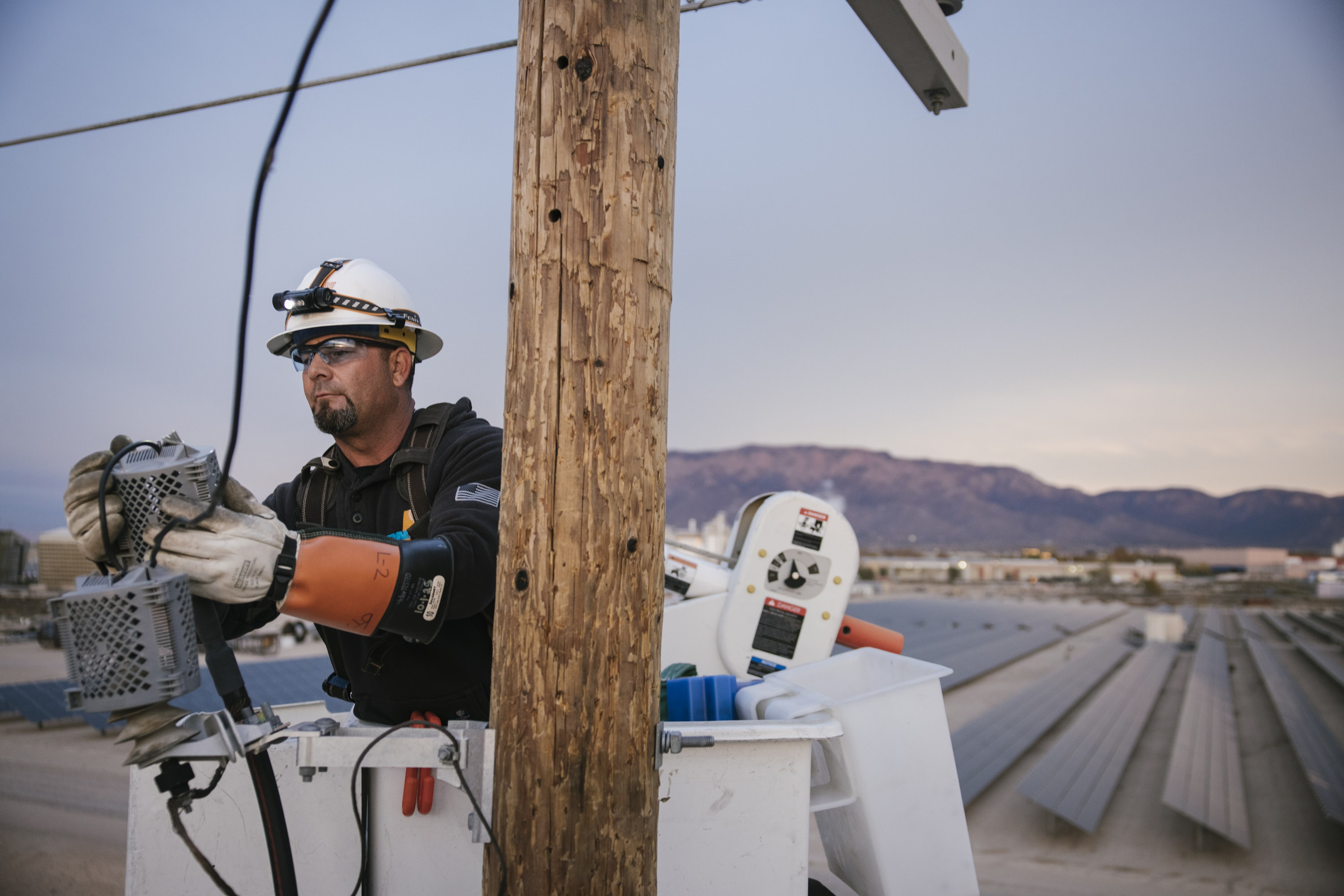 Electric utility worker with safety gear working on equipment from a bucket truck near solar panels under a clear sky.