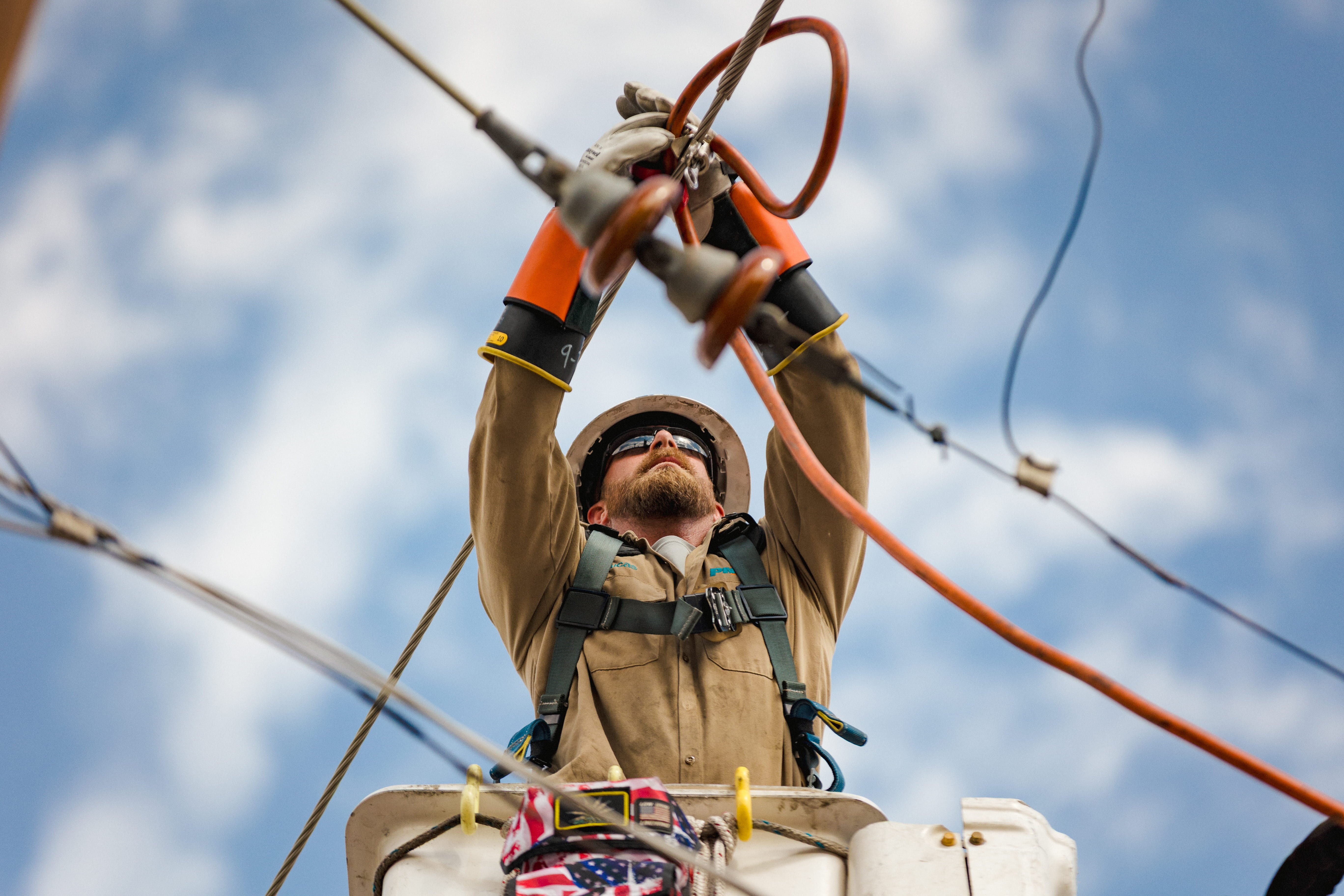 Utility worker wearing safety gear and gloves working on power lines from an elevated bucket against a blue sky.