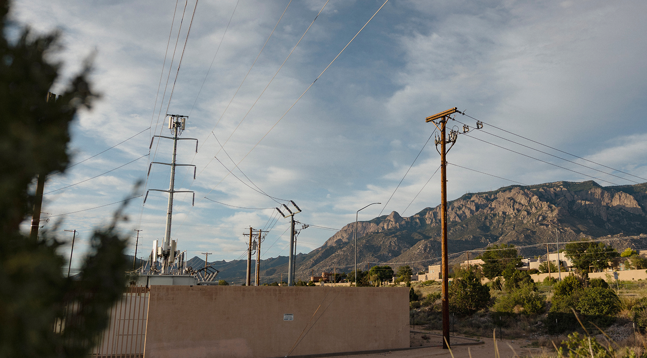Electrical utility poles and power lines against a blue sky with clouds and a mountain range in the background.