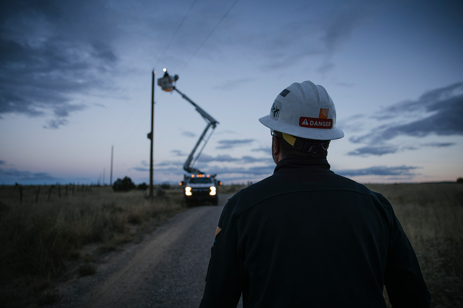 Worker wearing a hard hat with a danger label observing a bucket truck on a rural road during dusk.