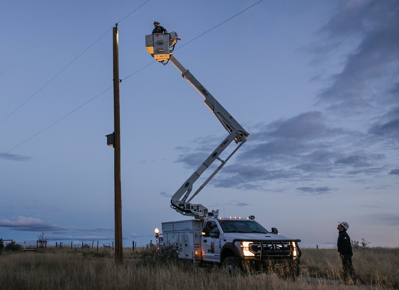 Utility worker in a bucket lift working on a power pole at dusk while another worker stands on the grass below.