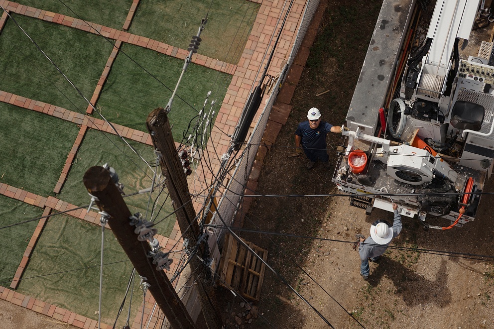 Two utility workers wearing hard hats working near a utility truck surrounded by power lines and poles.