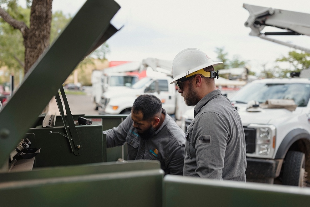 Two workers in gray uniforms inspecting equipment in the back of a truck, one wearing a white hard hat and sunglasses.