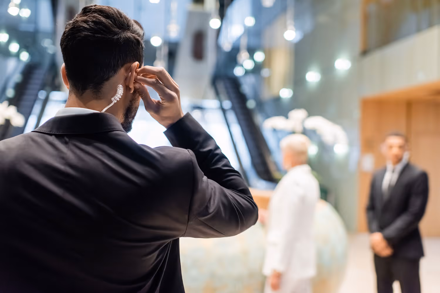 Security guard adjusting earpiece inside a modern building lobby with two other guards in the background.