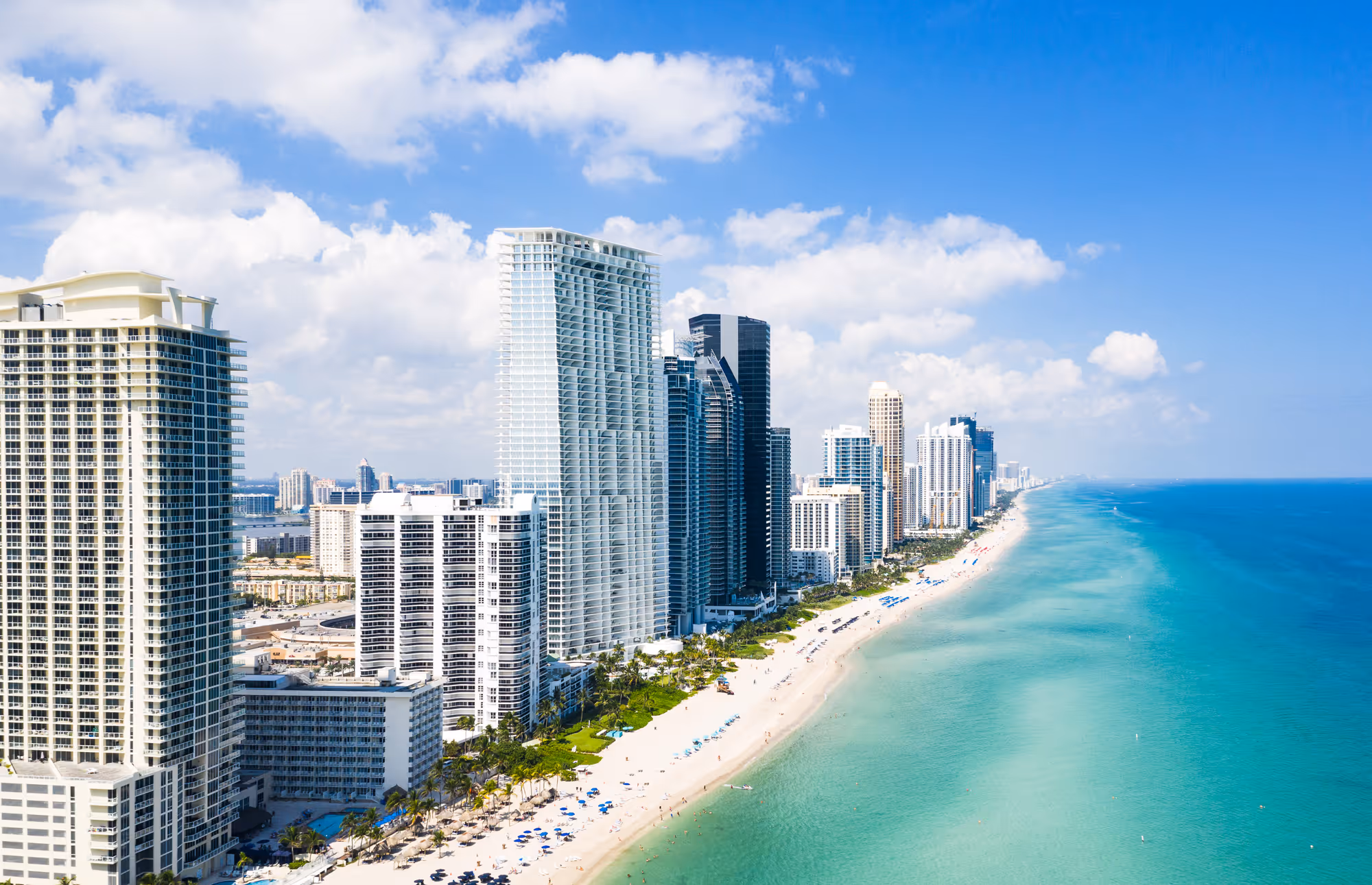 Aerial view of high-rise buildings in Miami lining a sandy beach with turquoise ocean under a partly cloudy blue sky.