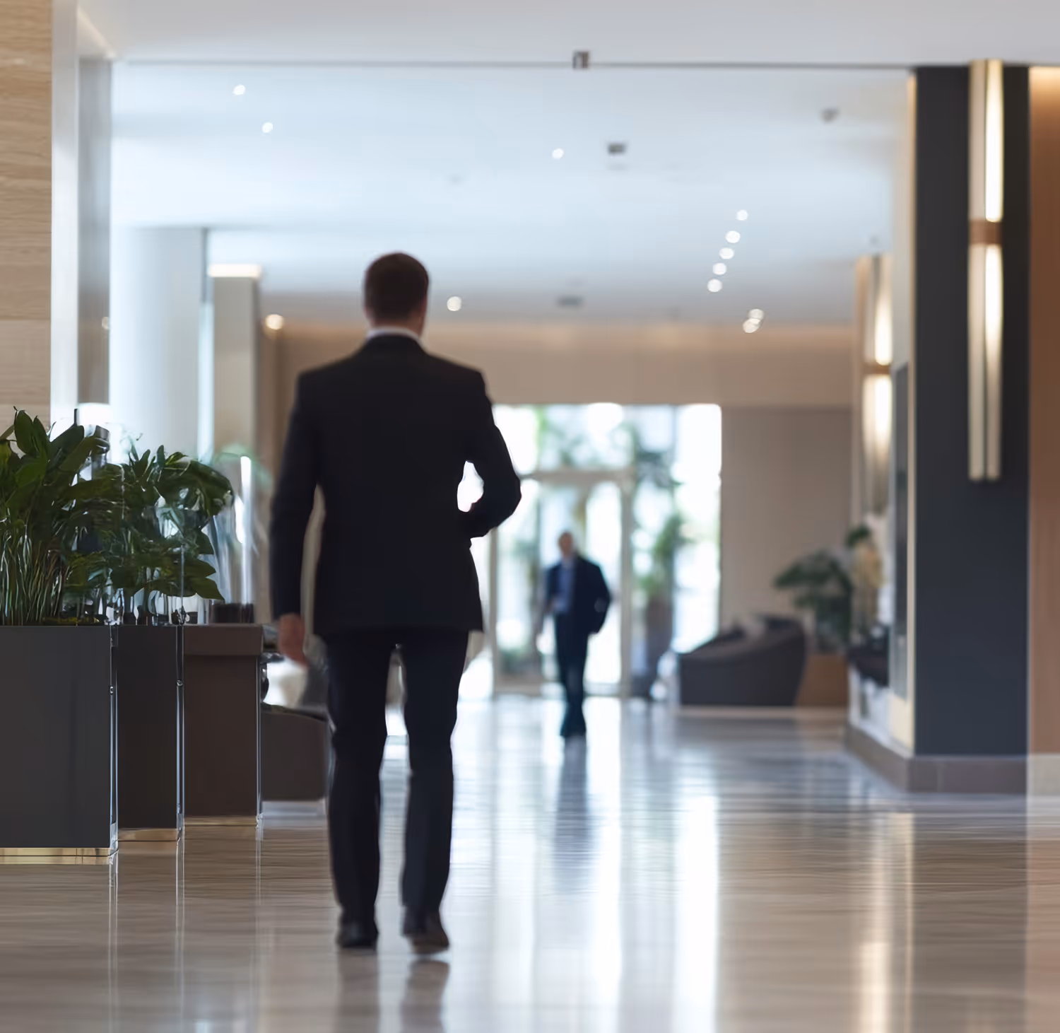 Man in a dark suit walking through a modern, spacious hotel lobby with polished floors and plants.