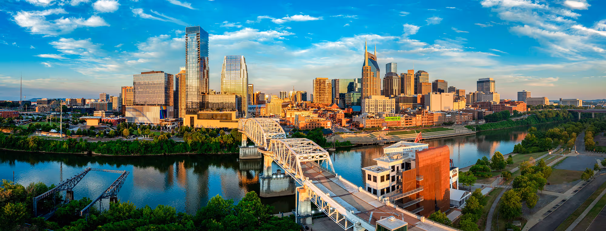 Panoramic view of Nashville skyline with modern buildings, a river, a bridge, and green trees under a partly cloudy blue sky.