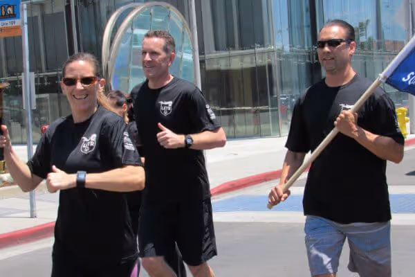 Three people jogging outside in black athletic shirts, one holding a flag, near a modern glass building.
