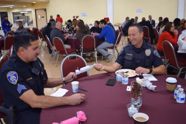 Two police officers sitting at a table sharing a Christmas cracker during a festive gathering with other people dining in the background.