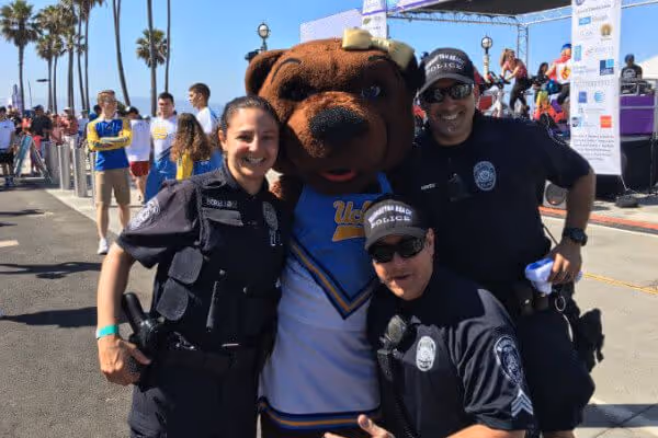 Three police officers posing with UCLA mascot bear at an outdoor event with palm trees and other people in the background.
