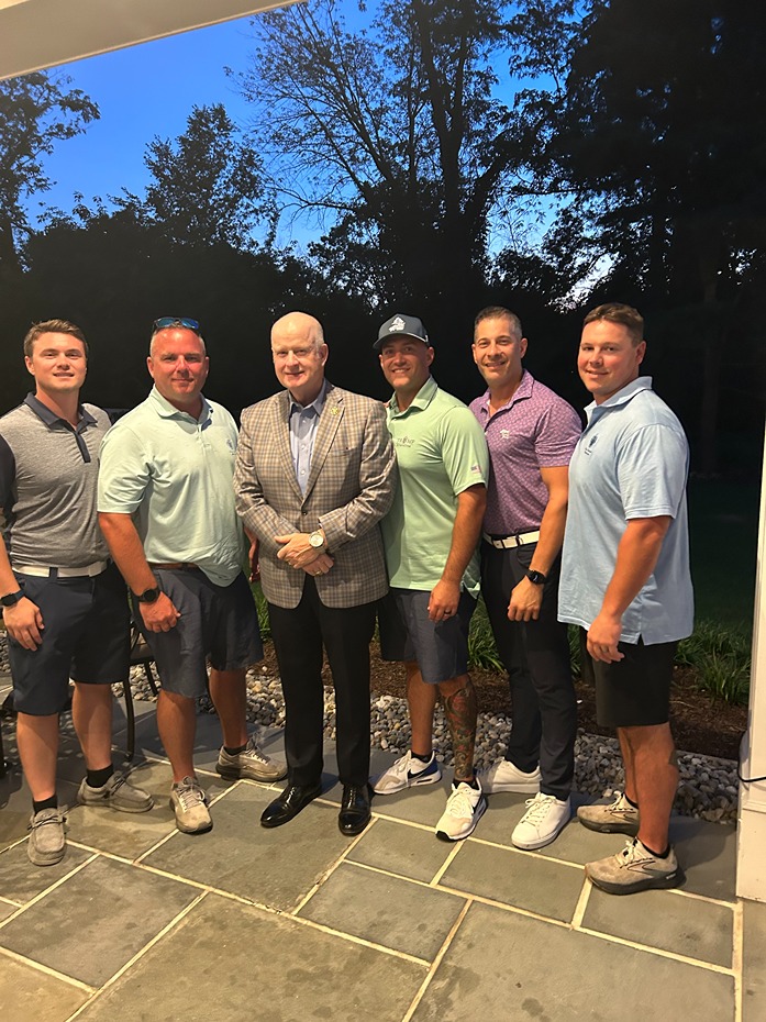 Six men standing together outdoors on a patio at dusk, five casually dressed and one in a suit jacket and dress shoes.