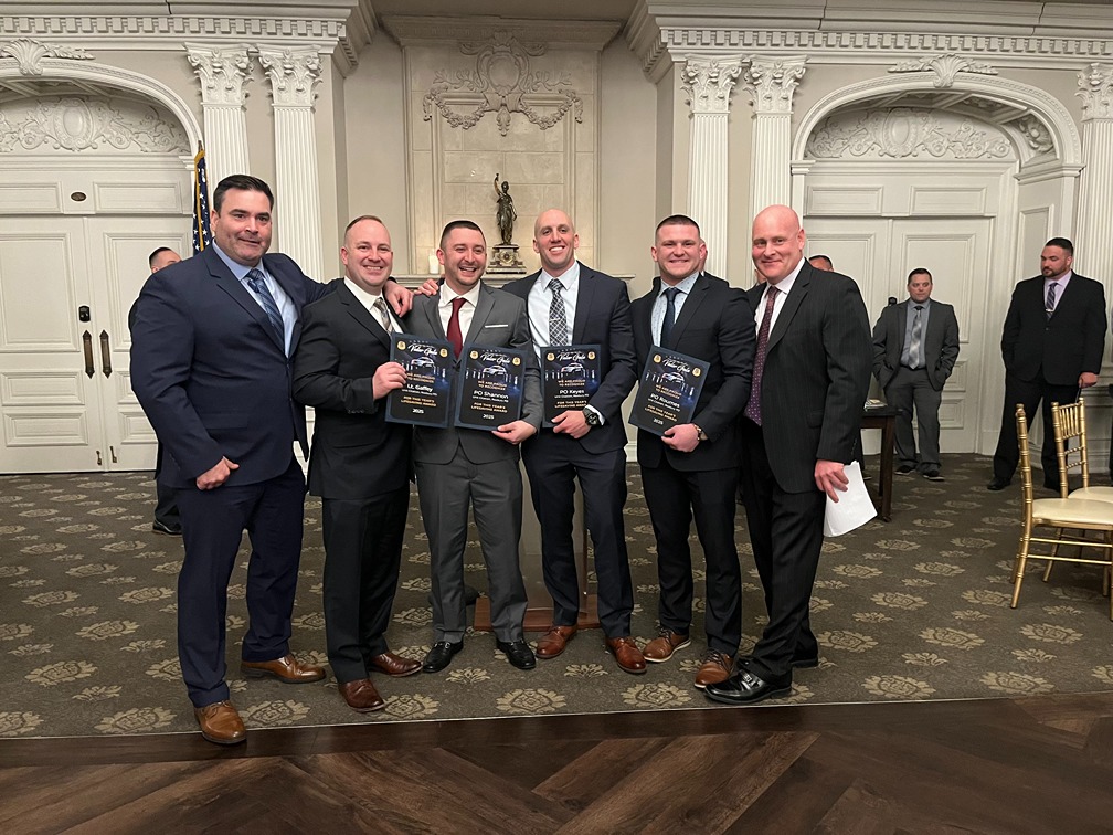 Group of six men dressed in suits standing indoors in front of ornate white walls and columns, four holding awards.