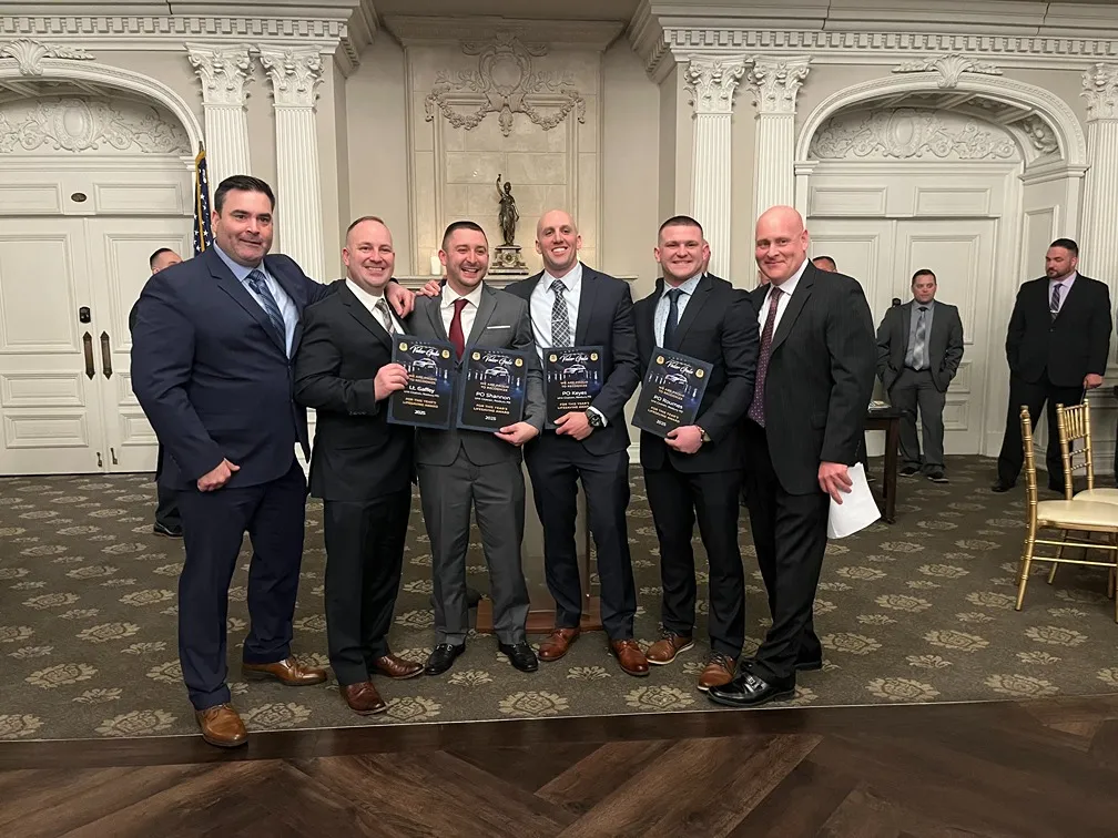 Group of six men dressed in suits standing indoors in front of ornate white walls and columns, four holding awards.