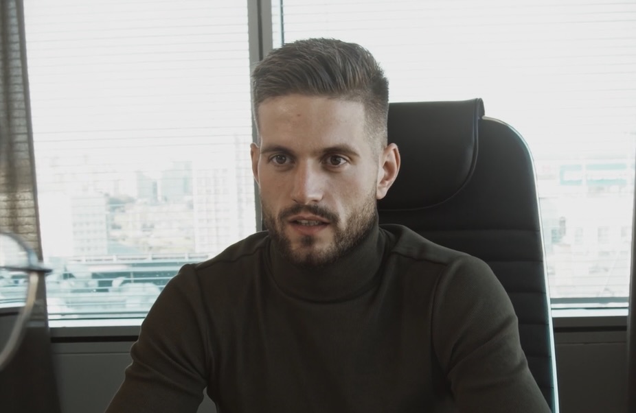 Man with short hair and beard wearing a dark turtleneck sitting in an office chair in front of a window.