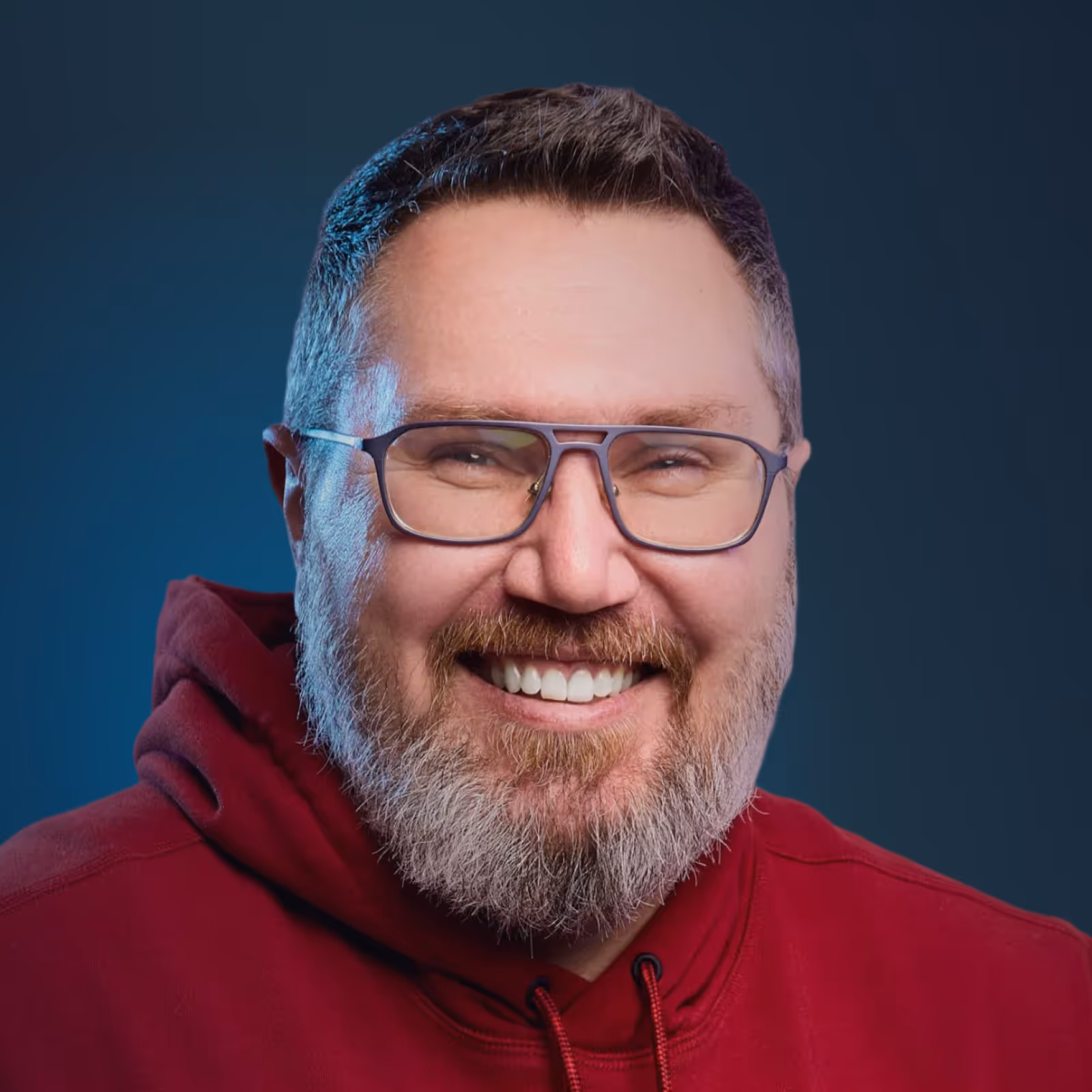 Smiling man with a short beard and glasses wearing a maroon hoodie against a dark blue background.