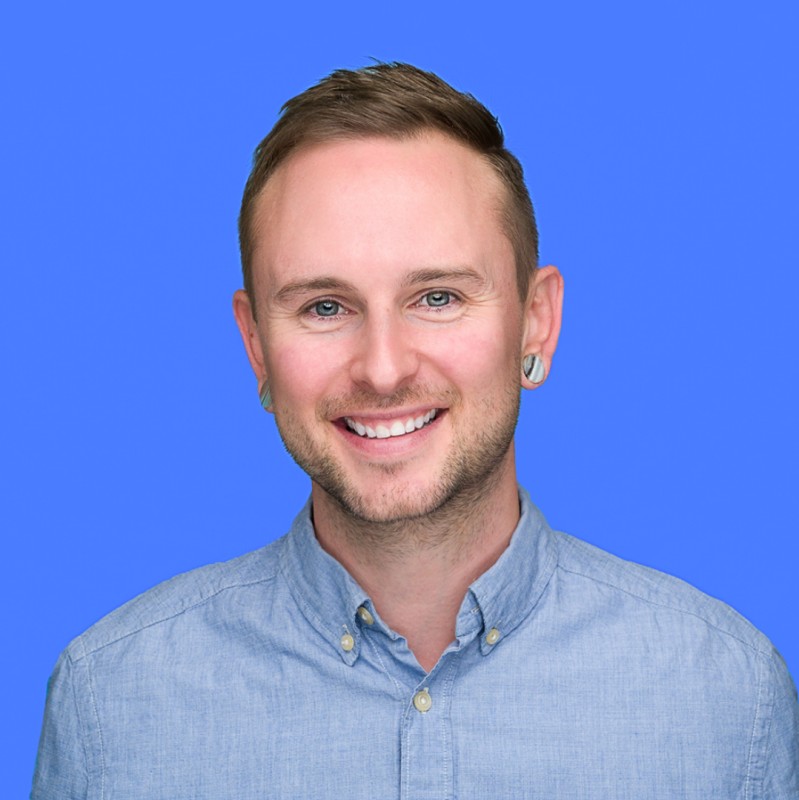 Smiling man with short hair wearing a light blue button-up shirt against a solid blue background.