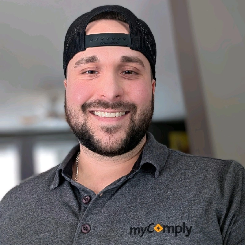 Smiling man with a beard wearing a black backwards cap and a grey polo shirt with the 'myComply' logo.