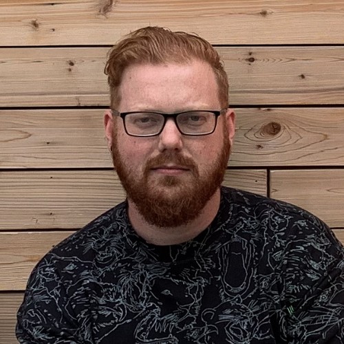 Man with red hair and beard wearing glasses and a black patterned shirt sitting in front of a wooden plank wall.