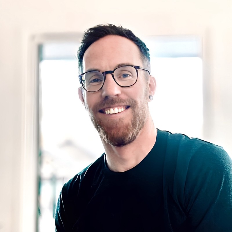 Smiling man with glasses, short hair, and beard wearing a black shirt in a bright room.