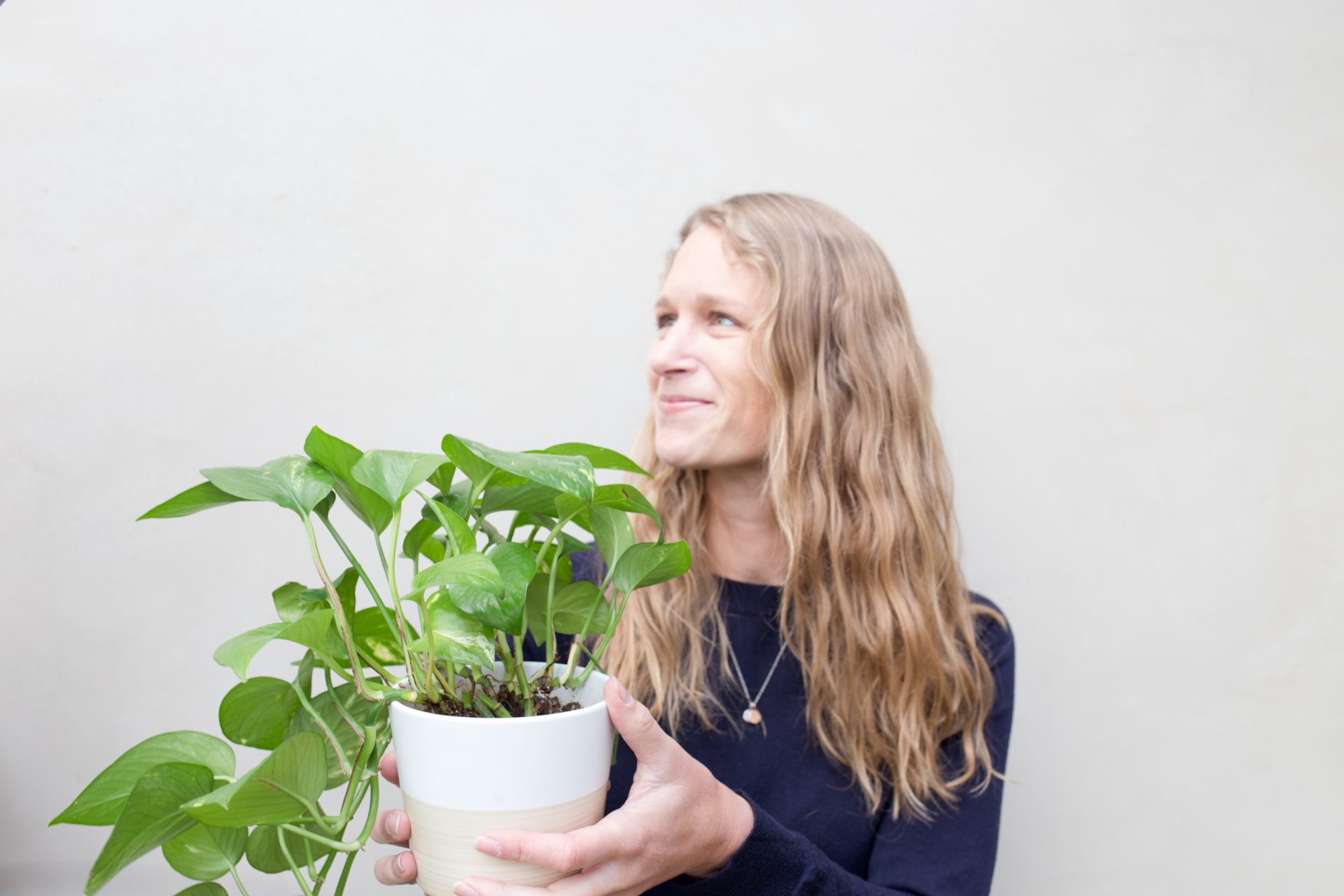 Woman with long wavy blonde hair wearing a dark sweater, holding a white plant pot with green leafy plant, smiling and looking to the side.