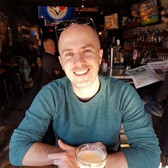 Smiling bald man wearing sunglasses on his head and a green sweater sitting at a bar with a glass of beer in front of him.