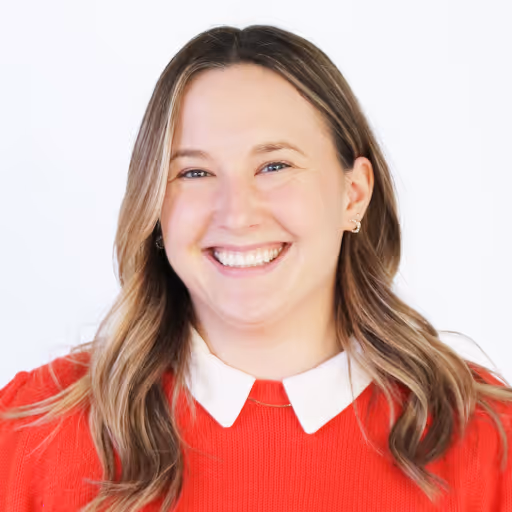 Smiling woman with long wavy hair wearing a red sweater with a white collar against a white background.