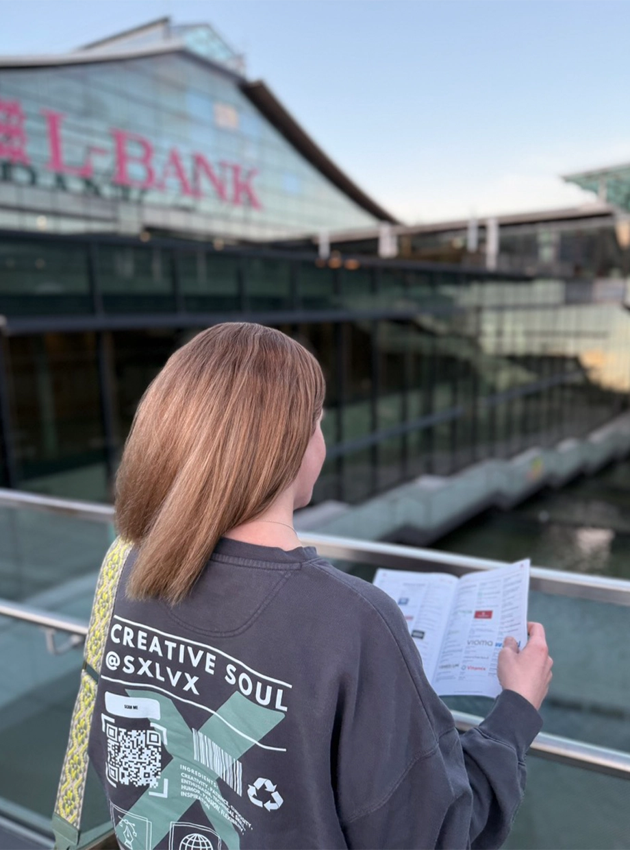 Two people sitting at a table with a laptop, one wearing an orange hoodie and the other a red beanie holding a mug, with pastries on the table.